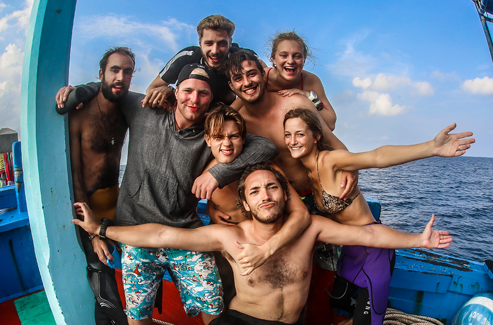 Happy group of scuba divers on boat deck after diving session, smiling and enjoying the ocean view, with blue sky and sea in the background.